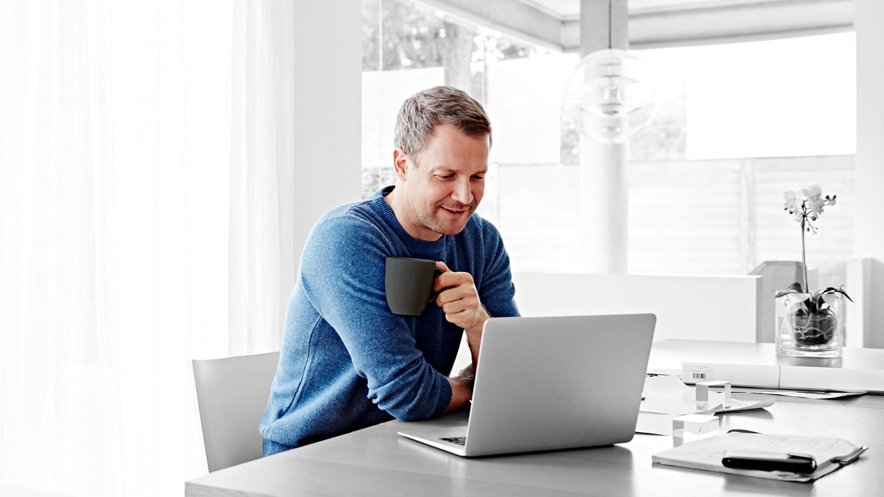A man using laptop and drinking a cup of coffee at living room; image used for HSBC Australia Coronavirus guidance.